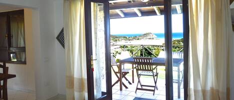 An open door leading to a patio with a table and chairs, a view of the sea, and a glimpse of the interior room.