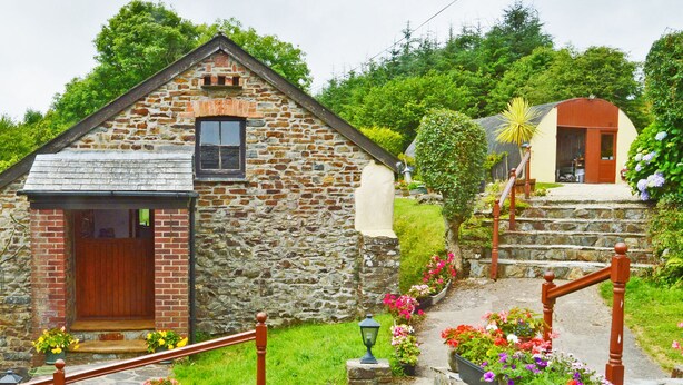 A stone building with a wooden door and steps leading up to it, surrounded by greenery and flowers.