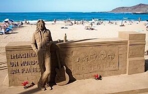 Eine Statue einer Person mit einer Gitarre am Strand, im Hintergrund ein klarer Himmel und das Meer.