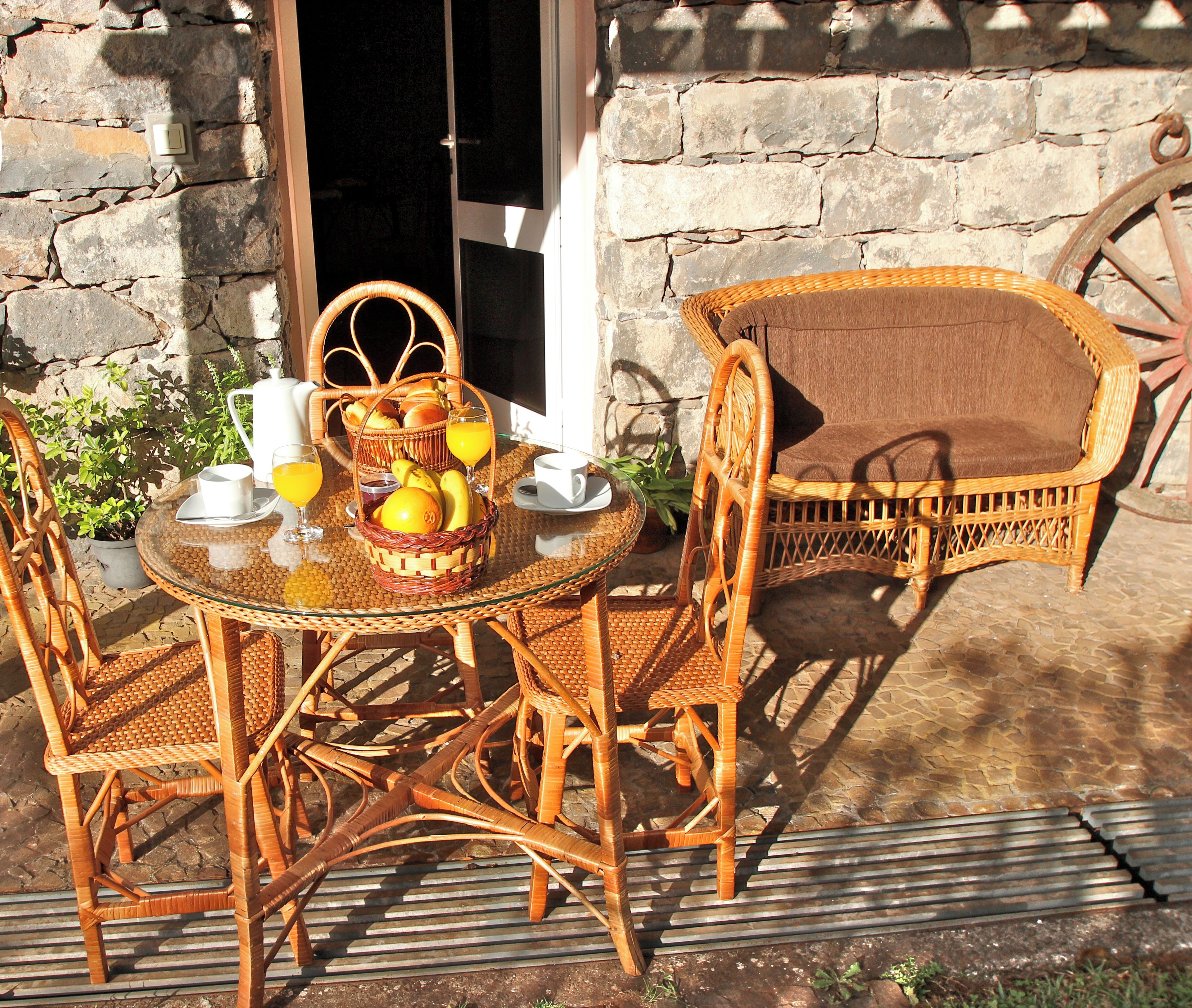A wicker table set with a basket of fruit, cups, and a pitcher, positioned outside a stone building with a sign reading "Clean & Safe."
