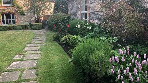 Un chemin de pierre mène à une maison traditionnelle à la façade en briques, dotée d’un jardin bien entretenu où poussent diverses plantes et fleurs.
