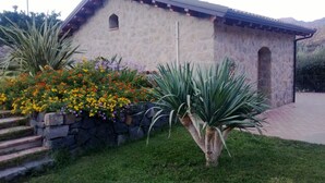 A stone building with a tiled roof, a stone staircase, and a garden with various flowers and a palm tree.