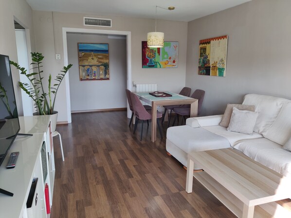 A living area with a sofa, a dining table with chairs, a TV, and a plant.