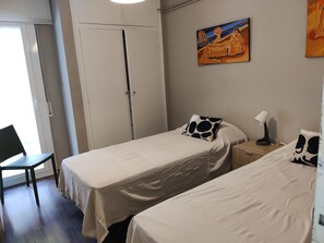 A room with two beds, a chair, a desk with a lamp, and a painting on the wall.