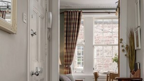 Un pasillo con piso de madera, una puerta blanca y un espejo con cuernos de ciervo. Se ve una ventana con cortina a cuadros y una pared de ladrillo a través de ella.