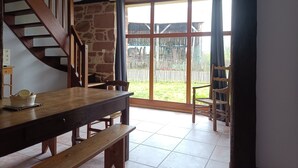 Een eetruimte met een houten tafel en banken, een trap en een groot raam met uitzicht op een omheinde tuin en gebouwen.