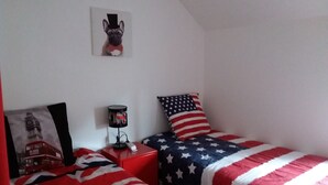 A bedroom with two beds, each covered with a red, white, and blue blanket. There is a lamp on a red nightstand, a picture of a dog on the wall, and a pillow with an American flag design. - In Marignac, house of 70m2, all comfort, with closed courtyard. (5 people) (Marignac)
