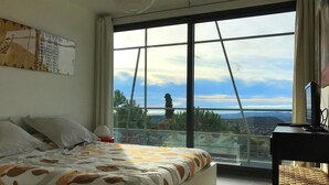 Une chambre avec un grand lit, un bureau avec un ordinateur et une vue sur le paysage urbain, visible à travers de grandes fenêtres.