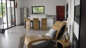 Un salon moderne avec un canapé aux lignes courbes, des chaises en bois et une cuisine en arrière-plan.