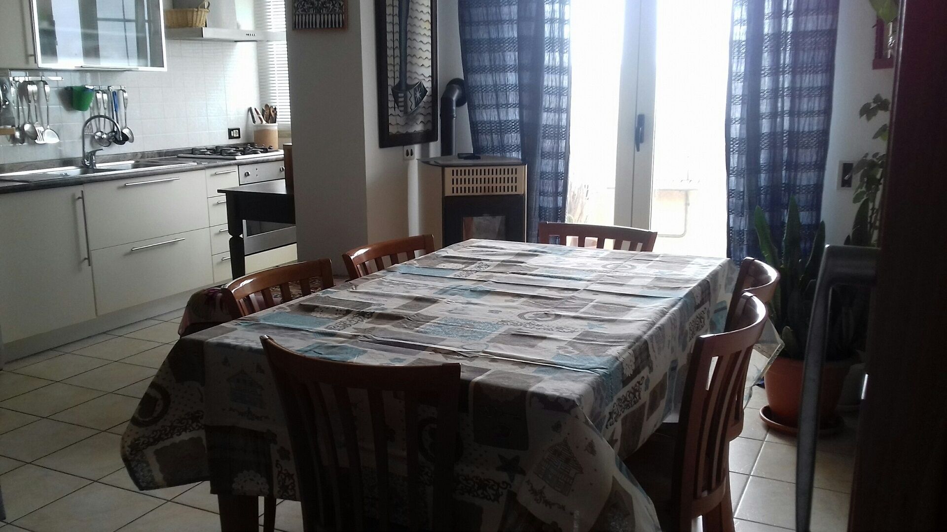 Un comedor con una mesa cubierta por un mantel estampado con motivos de periódico, rodeado de sillas de madera. Se ve una cocina con armarios blancos y una estufa al fondo.