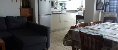 Un espacio habitable compacto con sofá, mesa de comedor y área de cocina, que incluye refrigerador, estufa y utensilios colgando sobre el fregadero.