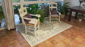 Una habitación con dos sillones blancos de mimbre con cojines morados, un tapete estampado y plantas en macetas.