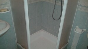 Een kleine, afgesloten douchecel met een witte vloer en betegelde wanden, een handdouche en een toilet met een papierrolhouder.