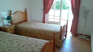 Een slaapkamer met twee houten bedden, een nachtkastje, een ventilator en een raam met gordijnen.
