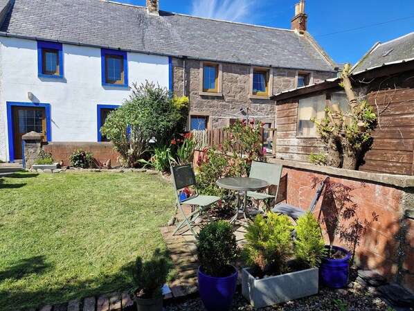 A traditional stone house with a well-maintained garden, a patio with a table and chairs, and a stone wall.