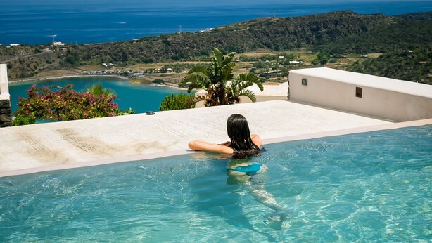 A woman relaxing in a clear blue infinity pool with a view of the ocean and hills. - RESIDENCE VENUS DREAM (Pantelleria)