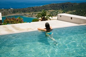 Eine Frau entspannt in einem klaren, blauen Infinity-Pool mit Blick auf das Meer und die Hügel.