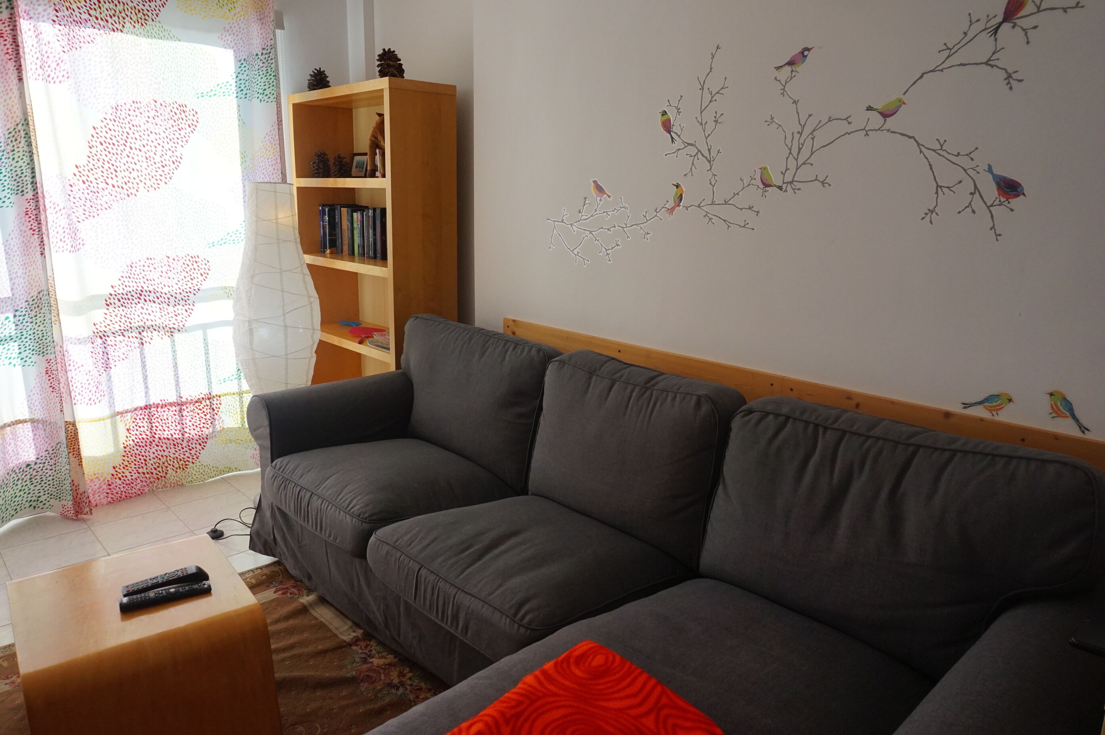 Uma sala de estar com um sofá cinzento, uma estante de madeira e um ornamento na parede com pássaros em galhos.