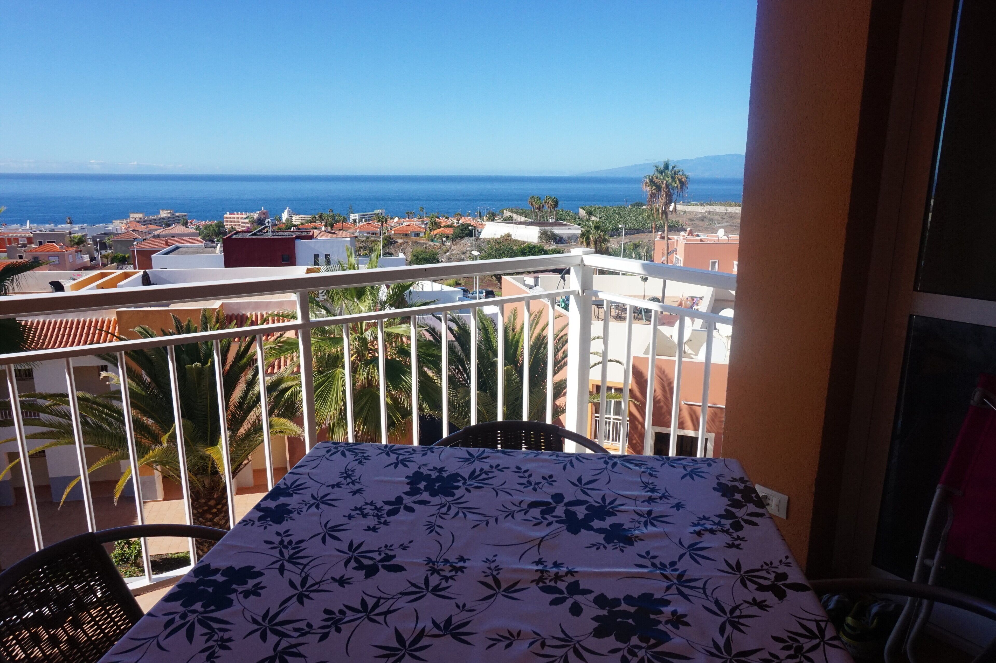 A balcony with a table and chairs, overlooking a coastal town and the sea.