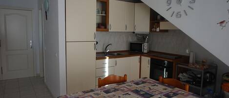 A compact kitchen with a microwave, sink, and wooden cabinets. A dining table with a quilted tablecloth is in the foreground.