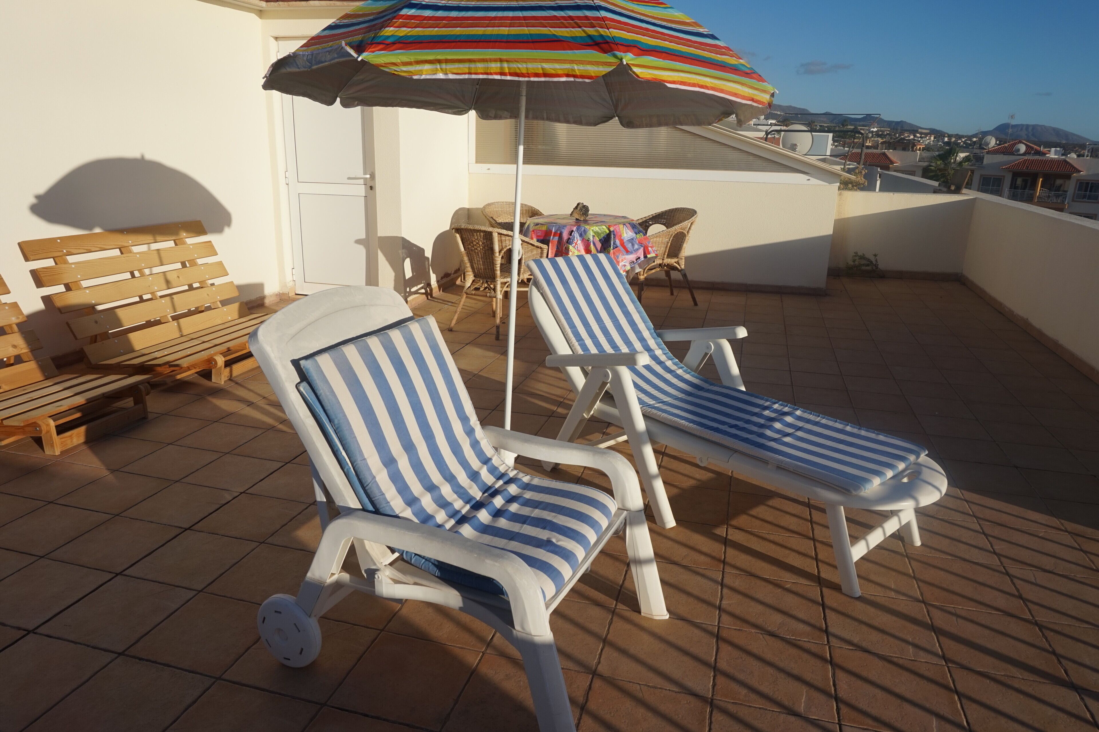 A rooftop terrace with two striped lounge chairs, a wooden bench, and a colorful umbrella.