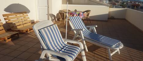 A rooftop terrace with two striped lounge chairs, a wooden bench, and a colorful umbrella.