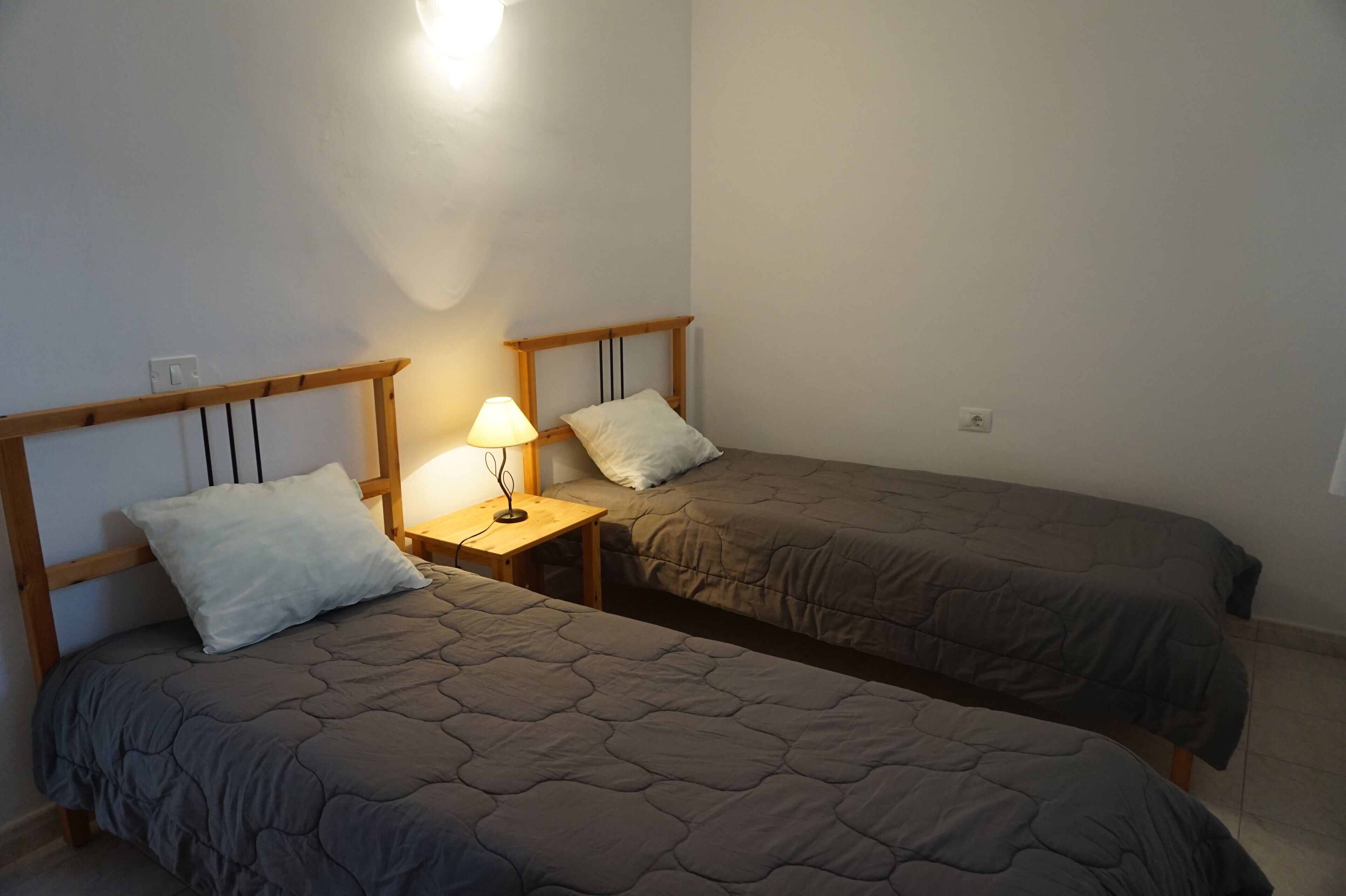 A room with two single beds, each with a wooden headboard and a bedside table with a lamp.