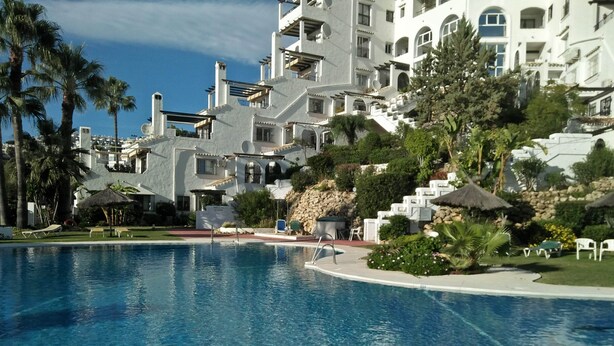 A swimming pool with a clear blue water surface, surrounded by palm trees and a white building with multiple balconies. - Apartment With Fabulous Sea Views, Excellent Facilities, Heated Indoor Pool (malaga)