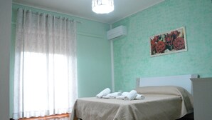 Una habitación con cama, ventana con cortinas, aire acondicionado y un cuadro con motivos florales en la pared.