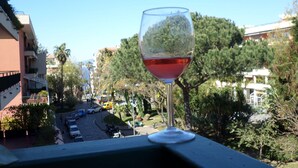 Vista despejada de una calle urbana con coches estacionados y árboles, una copa de vino tinto sobre una barandilla de balcón y edificios al fondo.