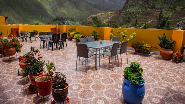 En terrasse med et bord og stole, omgivet af krukker med planter, med udsigt over et bjerglandskab.