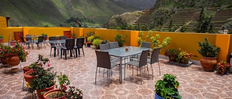 A terrace with a table and chairs, surrounded by potted plants, overlooking a mountainous landscape.