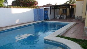Une piscine avec un bord carrelé, un patio couvert avec des chaises et une petite porte.