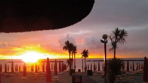 Une plage bordée de rangées de chaises longues et de parasols, de palmiers, et un coucher de soleil sur l’eau.