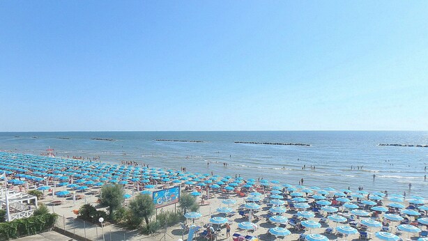 Une plage avec de nombreux parasols bleus et des chaises longues, un ciel dégagé et une mer calme.