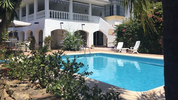 A poolside view of a white building with a balcony, surrounded by greenery and a clear blue pool. - Lovely Andalucian villa in a quiet, green neighborhood (Estepona)