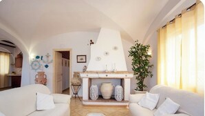 A living room with two white sofas, a fireplace with decorative vases, a potted plant, and a small table with a remote control. - Villa with private pool, large lawn, all with sea view. (Orosei)