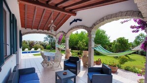 A covered patio with wicker furniture, a hammock, and a view of a garden and pool. - Villa with private pool, large lawn, all with sea view. (Orosei)