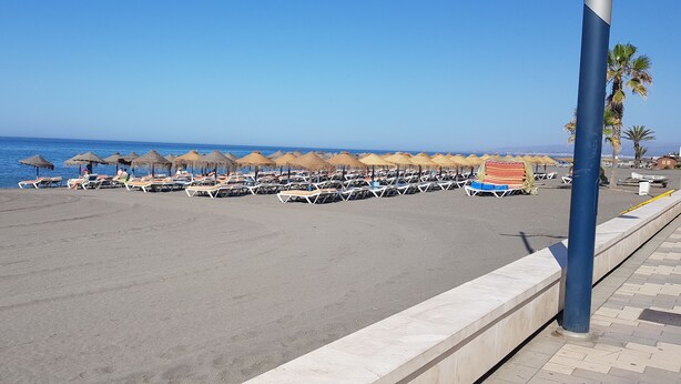 Een strand met rijen ligbedden en rieten parasols, een helderblauwe lucht en de zee op de achtergrond.