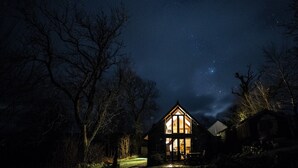 Una casa con un amplio ventanal central y techo inclinado, rodeada de árboles y un cielo nocturno estrellado.