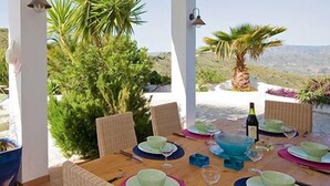 Een tafel voor vier met borden, kommen en een fles, omringd door rieten stoelen, met uitzicht op groen en heuvels.