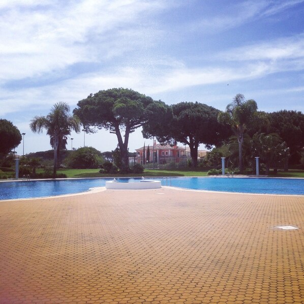 A swimming pool with a tiled deck, surrounded by trees and a building in the background. - Lovely 2 bedroom apartment with air con and excellent communal pool & bar (Fonte Santa)