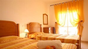 A hotel room with two beds, a wooden headboard, a bedside table with a lamp, and a window with yellow curtains. - Lovely 2 bedroom apartment with air con and excellent communal pool & bar (Fonte Santa)