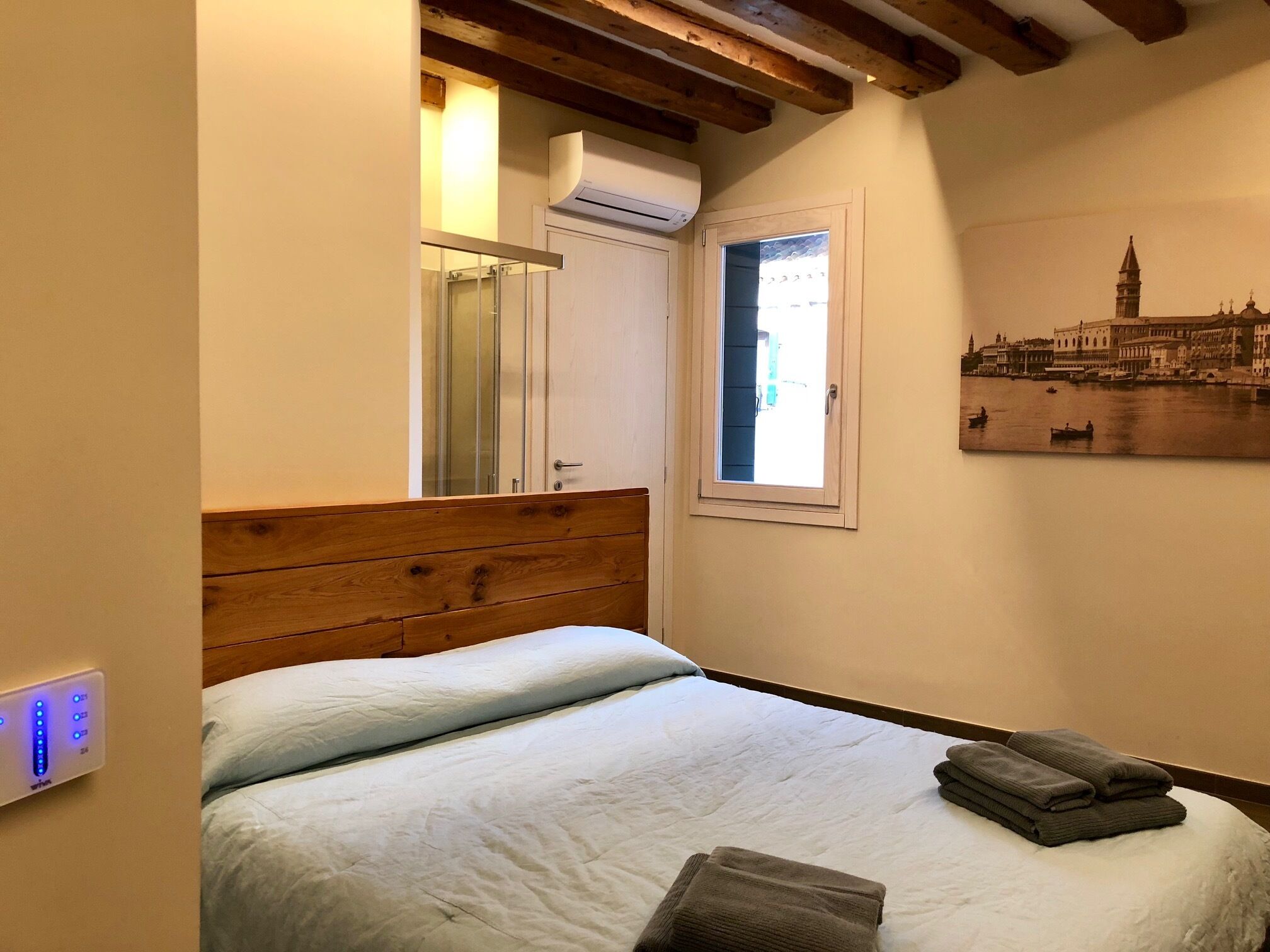 A bedroom with a wooden headboard, a bed with white bedding, a window with a view of a cityscape, and a wall-mounted air conditioning unit.