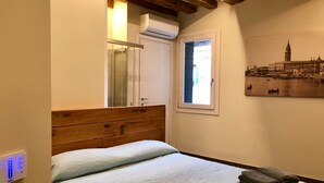 Un dormitorio con cabecera de madera, cama con ropa de cama blanca, ventana con vista a un paisaje urbano y un aire acondicionado de pared.