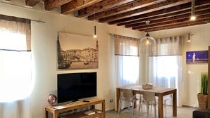 Una sala de estar con televisor, muebles de madera, una mesa de comedor con sillas y un ventanal con cortinas.