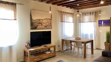 A living room with a TV, wooden furniture, a dining table with chairs, and a large window with curtains.