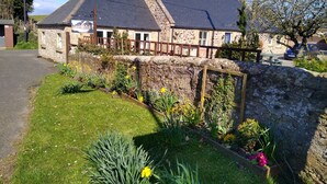 A stone building with a sloped roof, a stone wall, and a garden with various plants and flowers.