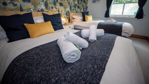 A hotel room with two beds, each with a dark grey bedspread, white linens, and yellow pillows. There are rolled towels on the beds and a window with curtains.