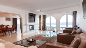 Amplia sala de estar con chimenea, comedor y un gran ventanal con vista al mar.